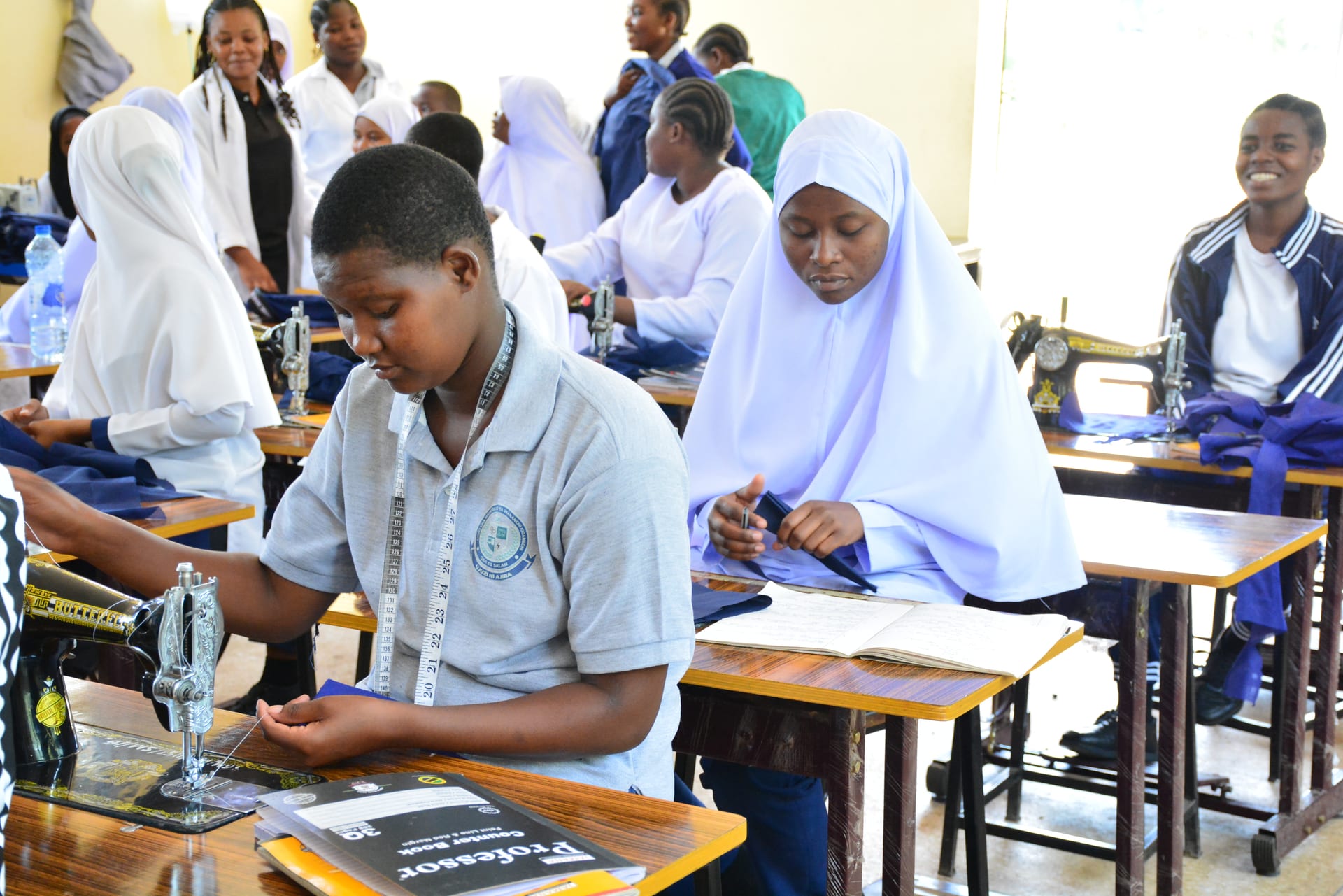 A full Folk Development College tailoring classroom at work: a student threading her Butterfly sewing machine in the foreground while classmates study, sew, and talk together behind her