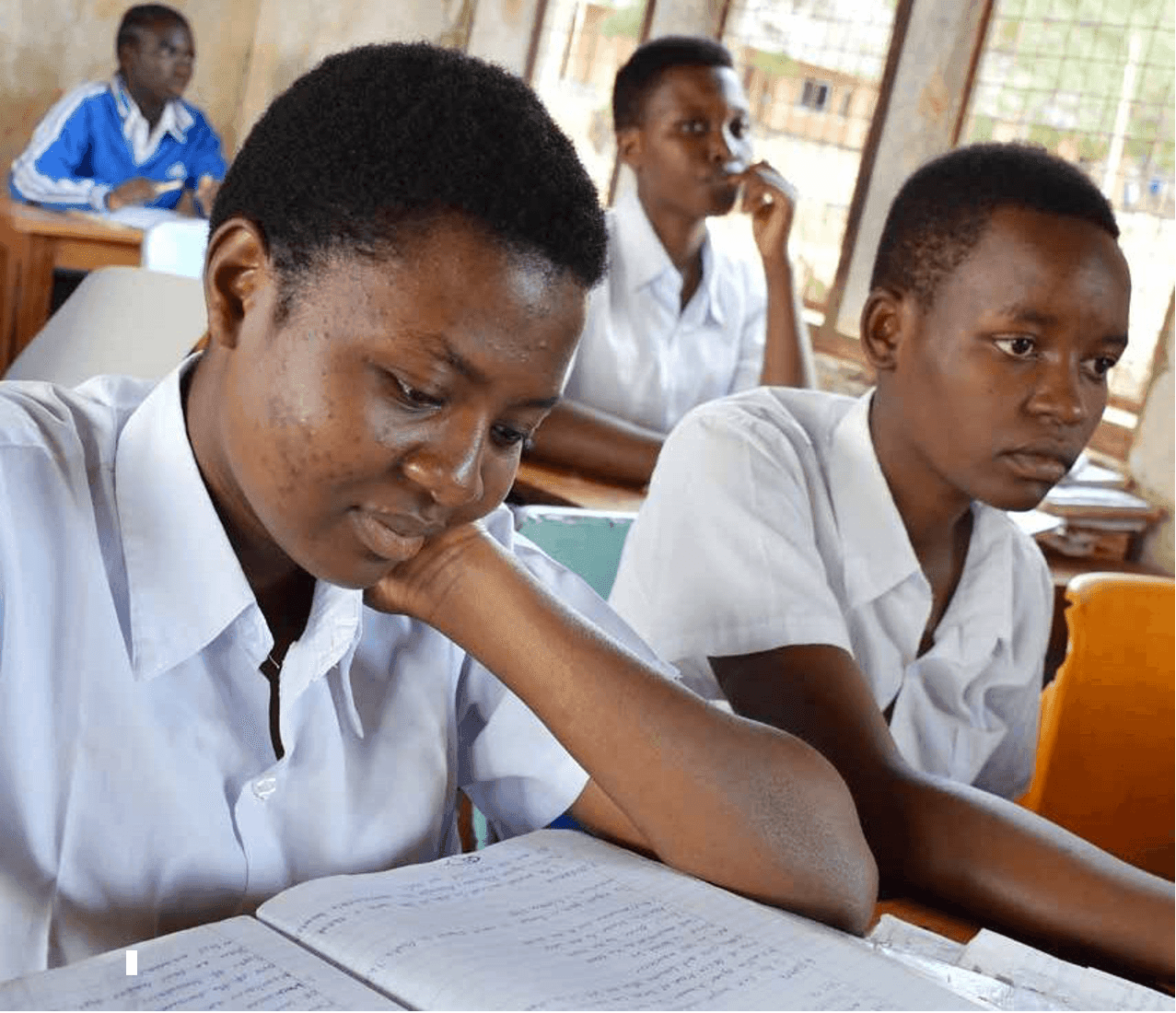 An FDC student in school uniform reading from her exercise book, hand under her chin, classmates focused around her in the classroom